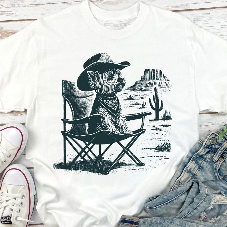 Pu&ograve; includere: T-shirt bianco con un'illustrazione in bianco e nero di un cane con cappello da cowboy e bandana, seduto su una sedia pieghevole. Lo sfondo raffigura un paesaggio desertico con un cactus e formazioni rocciose. Abbigliamento casual.