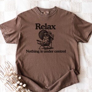 May include: Brown t-shirt with the word "Relax" above an illustration of a cat wearing sunglasses and a hat, sitting in a beach chair. The text "Nothing is under control" is below the illustration.