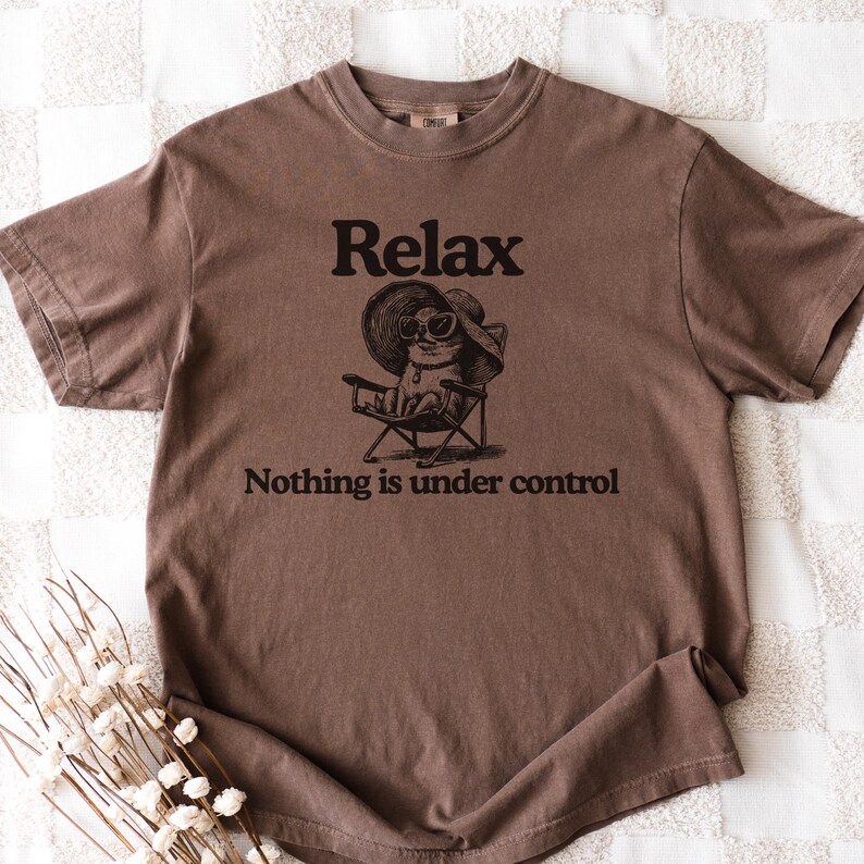 May include: Brown t-shirt with the word "Relax" above an illustration of a cat wearing sunglasses and a hat, sitting in a beach chair. The text "Nothing is under control" is below the illustration.