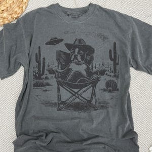 May include: Dark gray t-shirt featuring a dog wearing a cowboy hat, seated in a folding chair in a desert scene with cacti and a UFO. The graphic is in black and white.