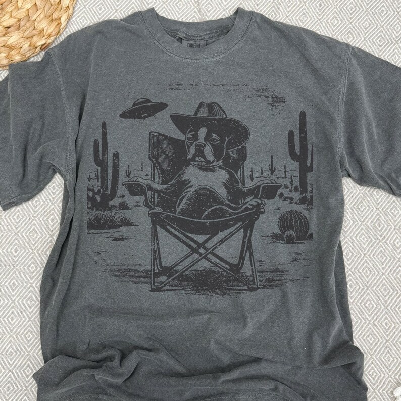 May include: Dark gray t-shirt featuring a dog wearing a cowboy hat, seated in a folding chair in a desert scene with cacti and a UFO. The graphic is in black and white.