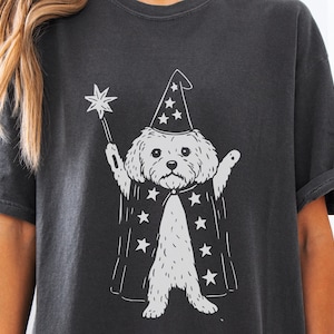 May include: Dark gray t-shirt featuring a white illustration of a dog dressed as a wizard. The dog wears a pointed hat and cape adorned with stars, holding a star-tipped wand. The shirt has short sleeves and a relaxed fit.