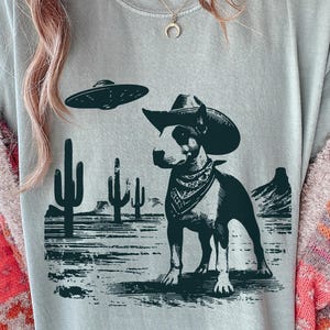 以下が含まれることがあります： カウボーイハットとバンダナを身に着けた犬のグラフィックが特徴のライトブルーのTシャツ。砂漠の風景にUFOが描かれています。犬は白黒です。背景にはサボテンと山があります。