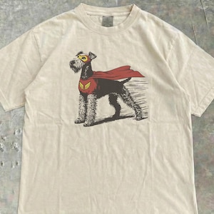 Puede incluir: Camiseta color crema con un gráfico de perro superhéroe. El perro está representado con una capa roja, una máscara amarilla y un emblema en el pecho. El gráfico está impreso en negro, rojo y amarillo.