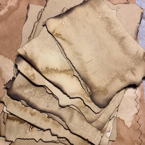Puede incluir: Una pila de hojas de papel marrón vintage con bordes irregulares y quemados. El papel tiene un aspecto desgastado y envejecido con manchas de agua visibles.