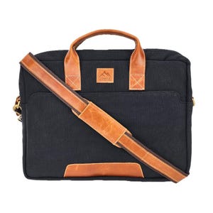 Könnte beinhalten: Schwarze Canvas-Aktentasche mit braunen Lederakzenten. Mit einem oberen Griff und einem abnehmbaren Schultergurt. Die Tasche hat eine rechteckige Form mit einer Vordertasche und einem kleinen Logo-Patch.