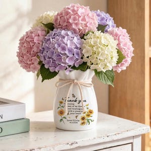 May include: A bouquet of colorful hydrangeas in a white vase. The flowers are various shades of pink, purple, and white. The vase has a tied bow and text that reads "Thank you" with sunflowers and butterflies. The vase is on a white surface.