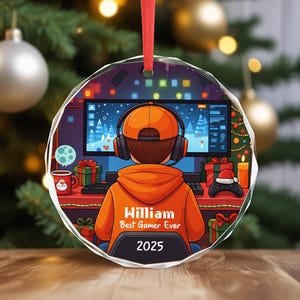 May include: A round, clear Christmas ornament with a red ribbon. The ornament features a gamer wearing an orange hoodie and headphones, sitting in front of a computer. Text on the hoodie reads "William Best Gamer Ever 2025".