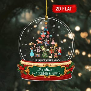 May include: A clear, round Christmas ornament with a gold string. The ornament features a snow globe design with a Nutcracker theme, including a Christmas tree, dancers, and toy soldiers. The text "THE NUTCRACKER 2025" and a name are visible.