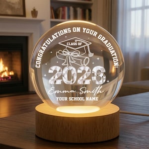 May include: A clear glass graduation sphere with the text "CONGRATULATIONS ON YOUR GRADUATION!" and "CLASS OF 2026". The sphere features a graduation cap and diploma design, and is personalized with a name and school name. It sits on a wooden base.