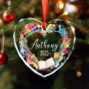 May include: Heart-shaped glass Christmas ornament with a red ribbon. The ornament features art supplies like paint tubes, brushes, and a watercolor palette. The name "Anthony" and the year "2025" are written on the ornament.