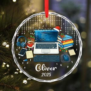 Pu&ograve; includere: Un ornamento natalizio rotondo e trasparente con un nastro rosso. L'ornamento presenta un laptop, tastiera, mouse, cuffie, tazza di caff&egrave; e libri. Il nome "Oliver" e l'anno "2025" sono stampati.