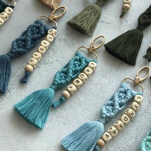 May include: A collection of macrame keychains in various colors, including teal, blue, green, and burgundy. Each keychain features a tassel, wooden beads with names, and a gold-colored clasp. The keychains are arranged on a light gray surface.