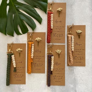 May include: A selection of handmade keychains in various colours, each featuring a name spelled out in wooden beads. The keychains are attached to cards with floral accents and the text "Will you be my..." and a date. Colours include red, orange, green, and brown.