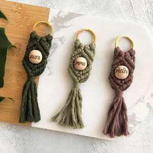 May include: Three macrame keychains in shades of green and brown, each with a gold-colored ring. The keychains feature a wooden bead with a name engraved on each: "Laura", "Emma", and "Shelby". The keychains have a tassel at the bottom.