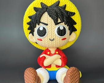 May include: A handmade, knitted Luffy doll from the anime series One Piece. The doll features a yellow hat, black hair, a red jacket, blue shorts, and brown shoes. The doll is sitting with its arms crossed and has large, expressive eyes.