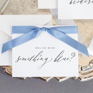 May include: White cards with light blue satin ribbon bows, and envelopes, arranged on a silver platter. The cards read "Will you be my bridesmaid?" and "Will you be my something blue?" A silver pen and white ribbon are also present.