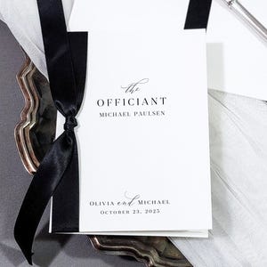 May include: A wedding program with the text "The Officiant Michael Paulsen" and the names "Olivia and Michael" with the date "October 23, 2025". The program is tied with a black ribbon and rests on a silver tray with a white tulle overlay.