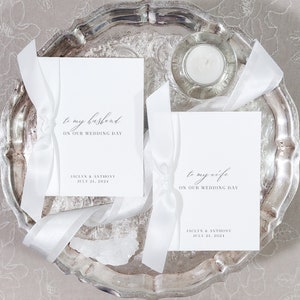 May include: Two white cards with gray text on a silver tray. The text on the cards reads "to my husband on our wedding day" and "to my wife on our wedding day". The cards also include the names "Jaclyn & Anthony" and the date "July 21, 2024".