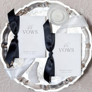 May include: Two white cardstock booklets with black ribbon tied around them. The top booklet says "his vows" and the bottom booklet says "her vows". Both booklets have the names "Kelsey & Riley" and the date "June 24, 2025" printed on them.