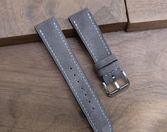 Pode incluir: Duas pulseiras de relógio cinza camurça com costuras brancas. Uma pulseira tem uma fivela prateada. As pulseiras são exibidas em uma superfície de madeira. As pulseiras têm aproximadamente 20 cm de comprimento.
