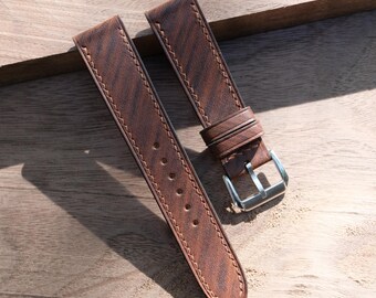 May include: A pair of brown leather watch straps with a textured grain pattern. The straps are stitched with a lighter brown thread and feature a silver-colored buckle. The watch bands are displayed on a wooden surface.