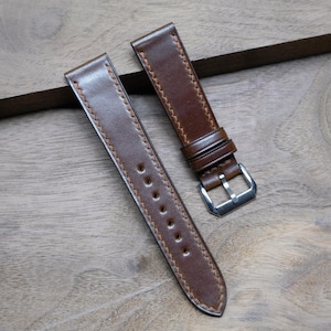 May include: Two brown leather watch straps with light brown stitching. One strap has a silver buckle. The straps are on a wooden surface. The leather has a smooth texture and a slight sheen.