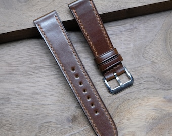 Könnte beinhalten: Zwei braune Leder-Uhrenarmbänder mit hellbraunen Nähten. Eines der Armbänder hat eine silberne Schnalle. Die Armbänder liegen auf einer Holzoberfläche. Das Leder hat eine glatte Textur und einen leichten Glanz.