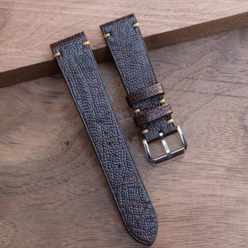 Puede incluir: Dos correas de reloj de cuero marr&oacute;n oscuro con una superficie texturizada y costuras beige. Una correa tiene una hebilla plateada. Las correas se muestran sobre una superficie de madera.