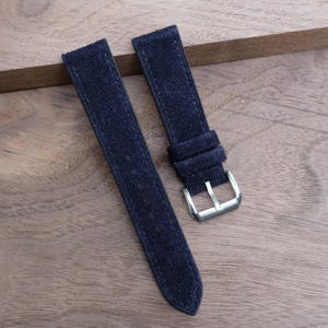 May include: Two navy blue suede watch straps with a silver-colored buckle. The straps are made of a soft, textured material and are designed to attach to a watch face. The straps are shown on a wooden surface.