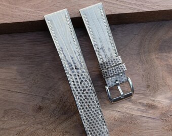 Könnte beinhalten: Zweiteiliges Uhrenarmband in hellgrauem, strukturiertem Muster mit silberfarbener Schnalle. Das Armband besteht aus einem reptilienartigen Material und ist mit weißem Faden genäht. Das Uhrenarmband wird auf einer Holzoberfläche präsentiert.