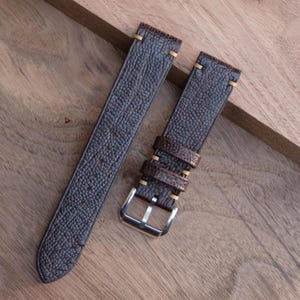 Puede incluir: Correa de reloj de dos piezas en cuero marr&oacute;n oscuro con acabado texturizado. La correa presenta costuras doradas y una hebilla plateada. El cuero tiene un aspecto granulado y se muestra sobre una superficie de madera.