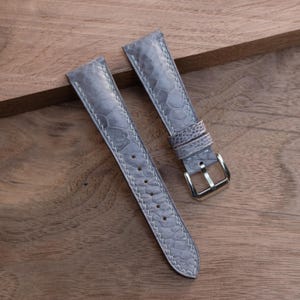 Puede incluir: Dos correas de reloj grises con un patrón texturizado similar al de un reptil y costuras blancas. Una correa tiene una hebilla plateada. Las correas se muestran sobre una superficie de madera.