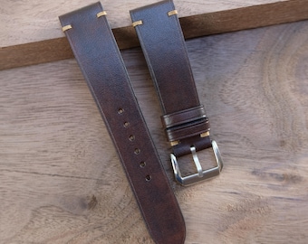 May include: Two dark brown leather watch straps with cream-colored stitching. One strap has a silver buckle. The straps are lying on a wooden surface.