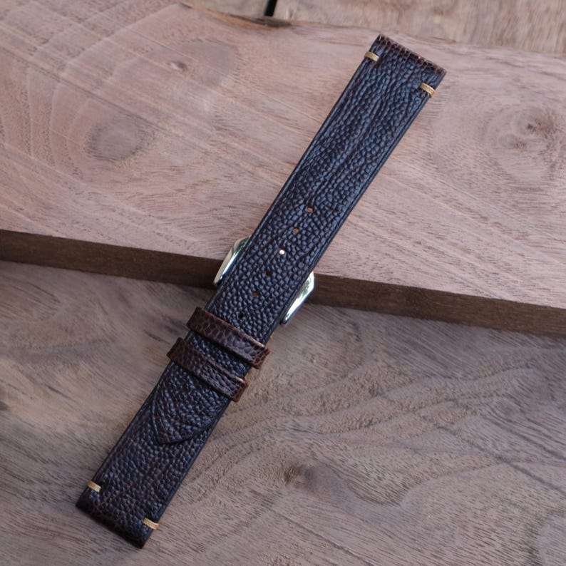 Puede incluir: Una correa de reloj de cuero marr&oacute;n oscuro con una superficie texturizada. La correa presenta una hebilla plateada y una presilla de cuero marr&oacute;n m&aacute;s peque&ntilde;a. Los extremos de la correa tienen costuras doradas.