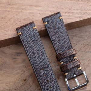 Puede incluir: Dos correas de reloj de cuero marr&oacute;n con superficie texturizada y costuras de color beige claro. Una correa tiene dos presillas y una hebilla plateada. Las correas se muestran sobre una superficie de madera.