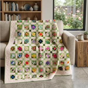 May include: A floral patchwork quilt draped over a beige sofa. The quilt features a grid pattern with squares of various colourful flower designs. The border is a light pink with a quilted pattern. The background includes a bookshelf and a window.