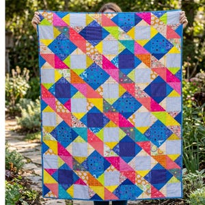 Puede incluir: Un edredón colorido con un patrón geométrico. Presenta cuadrados y triángulos en tonos de azul, rosa, amarillo, naranja y gris. El edredón se muestra sobre un fondo de follaje verde.