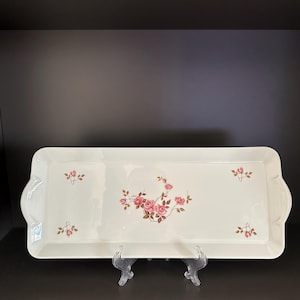 Può includere: Un vassoio rettangolare bianco sporco con un motivo floreale a rose. Il vassoio ha un bordo smerlato ed è sostenuto da un supporto trasparente. Le rose sono rosa e marroni.