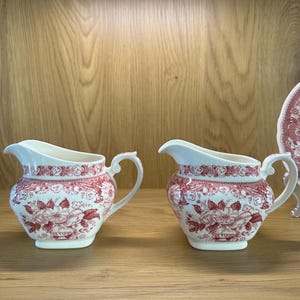 May include: Two matching cream jugs with a white base and a red floral pattern. Each jug has a curved handle and a spout. The jugs are displayed on a wooden shelf.