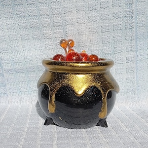 May include: A black cauldron with gold glitter accents and a gold rim, filled with red and orange glass beads. The cauldron has three small feet and a dripping gold design.