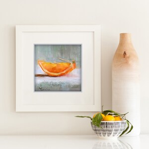 May include: A framed painting of an orange slice, with a white frame. The painting is on a white surface, with a decorative vase and a bowl of lemons nearby. The painting has a signature in the bottom left corner.