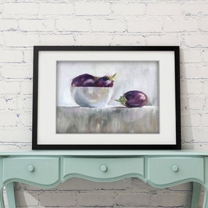 May include: A framed still life painting featuring a white bowl filled with purple eggplants and one eggplant resting beside the bowl. The painting is set against a neutral background and framed in black. The artwork is displayed on a light blue table.