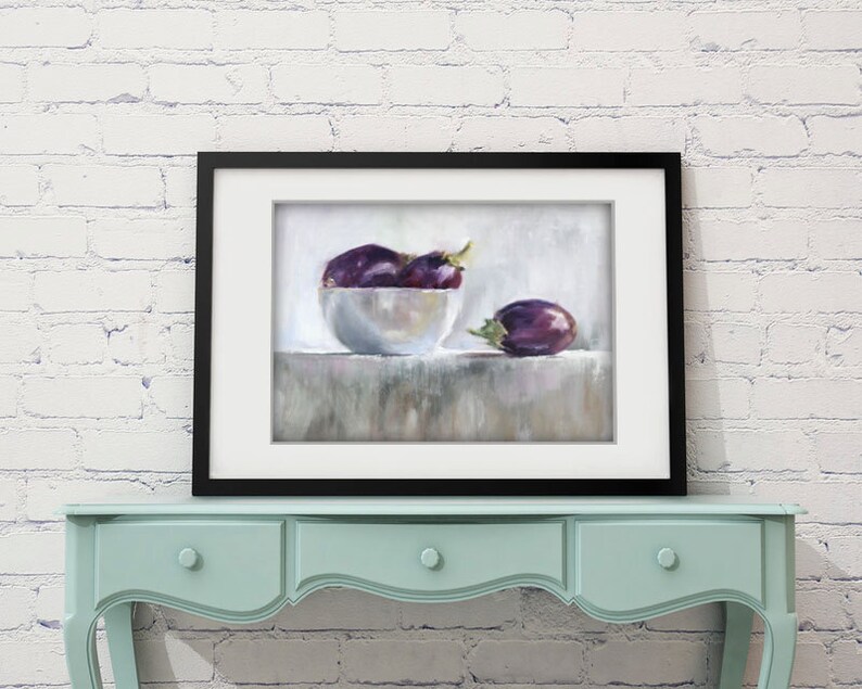 May include: A framed still life painting featuring a white bowl filled with purple eggplants and one eggplant resting beside the bowl. The painting is set against a neutral background and framed in black. The artwork is displayed on a light blue table.
