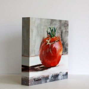 Puede incluir: Pintura al &oacute;leo de un tomate rojo maduro con tallos verdes. La obra de arte est&aacute; sobre un peque&ntilde;o lienzo rectangular, mostrando la textura del tomate y el juego de luces y sombras. El fondo es gris.