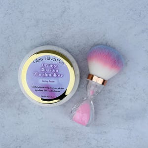 May include: A round, off-white container with a gold rim and a purple label that reads "Glow Haven Co. Orange Blossom Marshmallow Dusting Powder." A makeup brush with a pink, white, and blue ombre effect sits next to it. The container is 3 oz.