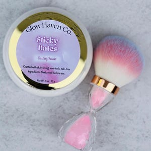 May include: A white container of "Sticky Dates" dusting powder with a gold rim and a makeup brush. The brush has a pink and blue ombre effect on the bristles and a clear hourglass-shaped handle. The container reads "Glow Haven Co."