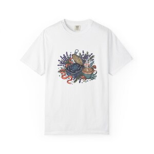 Puede incluir: Camiseta blanca con una ilustración colorida de una lata de sardinas, una taza de café y ramitas de lavanda. Una cinta roja se arremolina alrededor del diseño. La camiseta tiene cuello redondo y mangas cortas.