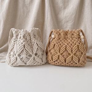 May include: Two drawstring bags crafted with macrame cord in off-white and tan colors. Each bag features a repeating diamond pattern, a braided shoulder strap, and a single wooden bead accent on the drawstring cord for a secure closure.