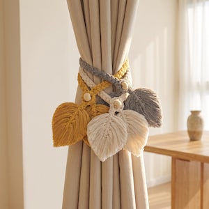 May include: Three macrame curtain tiebacks featuring braided cords and leaf-shaped tassels in mustard yellow, cream, and grey. Each tieback is accented with a single round wooden bead where the braided cord meets the fringed leaf design.